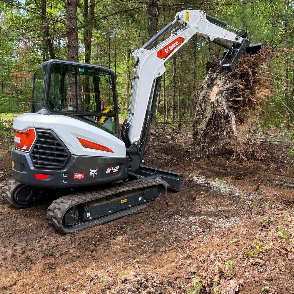 Compact excavator removing tree roots in wooded area with forest background.