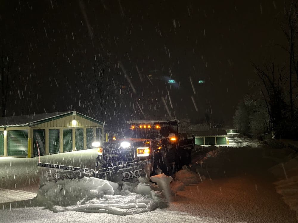 Snow plow clearing heavy snowfall at night, illuminated by surrounding lights.