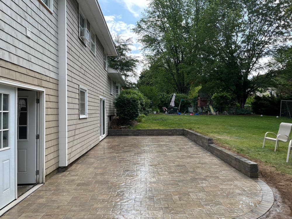 Patio area in backyard with stone tiles, lawn, and trees, featuring a house in the background.