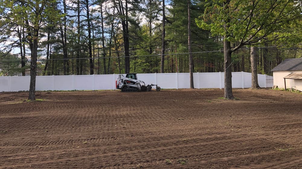Bobcat machinery preparing a cleared, leveled yard surrounded by trees and a white fence.