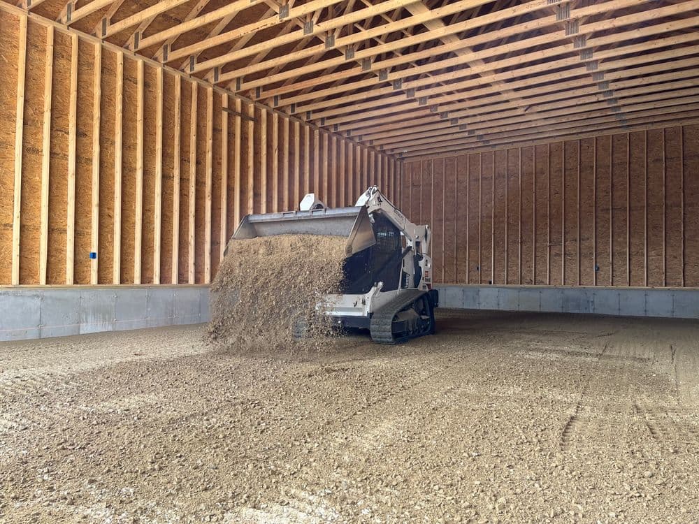 Skid steer loader dumping gravel inside a newly constructed barn with wooden beams.