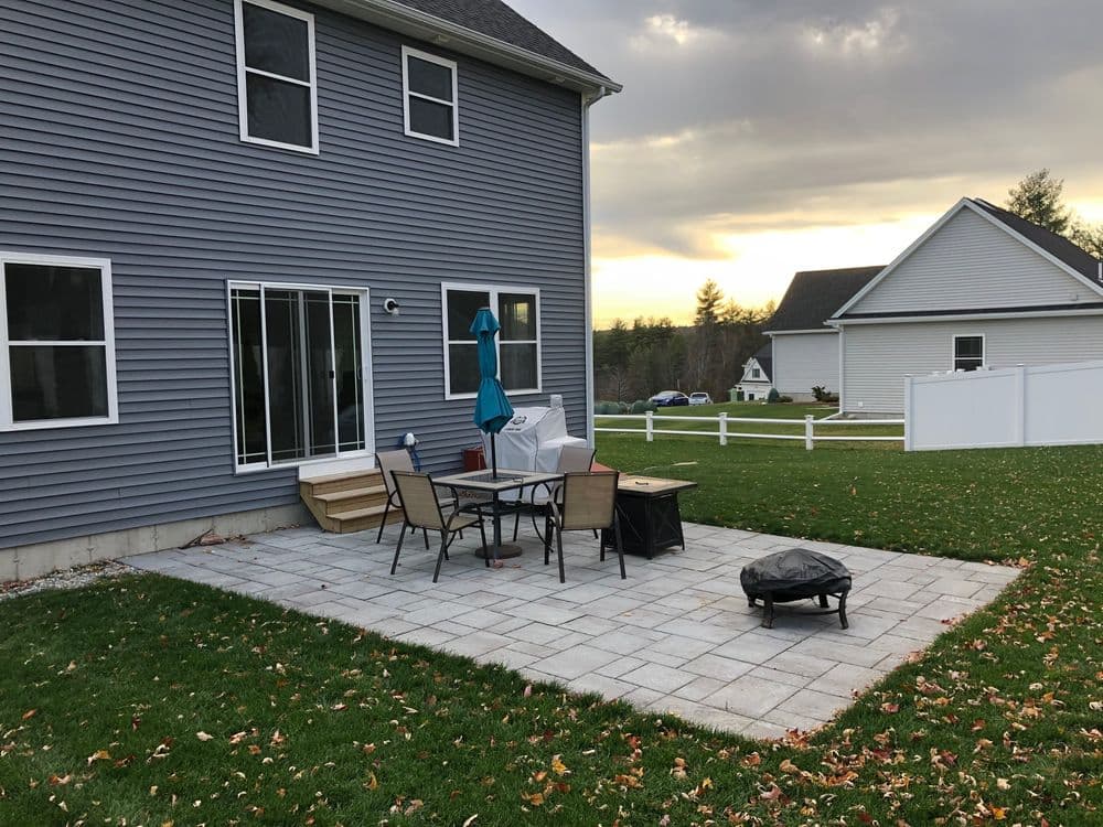 Patio area with table, chairs, and grill beside a gray house, surrounded by green lawn.